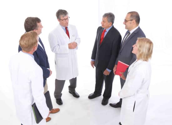 A group of physicians stand in a semicircle talking against a white background.