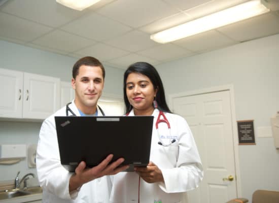 A male and female physician smile as they study a patient's chart on a laptop.